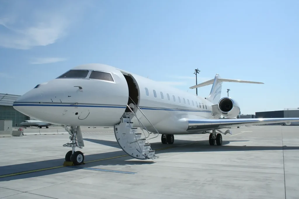 This image shows a Bombardier Global 6000 business jet from a front-side angle, parked on the tarmac with its stairs extended, ready for boarding. The sleek white exterior and the open door emphasize its readiness for passengers, with clear skies and an airport setting in the background.
